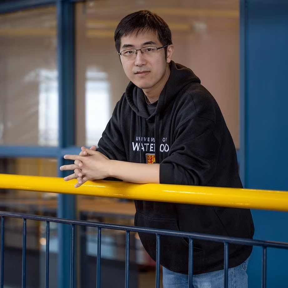 An Asian man donning a black UWaterloo sweatshirt posing aganist a yellow railing aganist a blue-painted wall and a large mirror