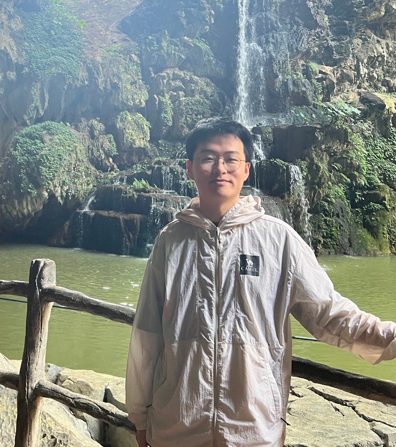 Asian man wearing a white rain jacket posing in front of a green-coloured lake, lush trees and a waterfall. 