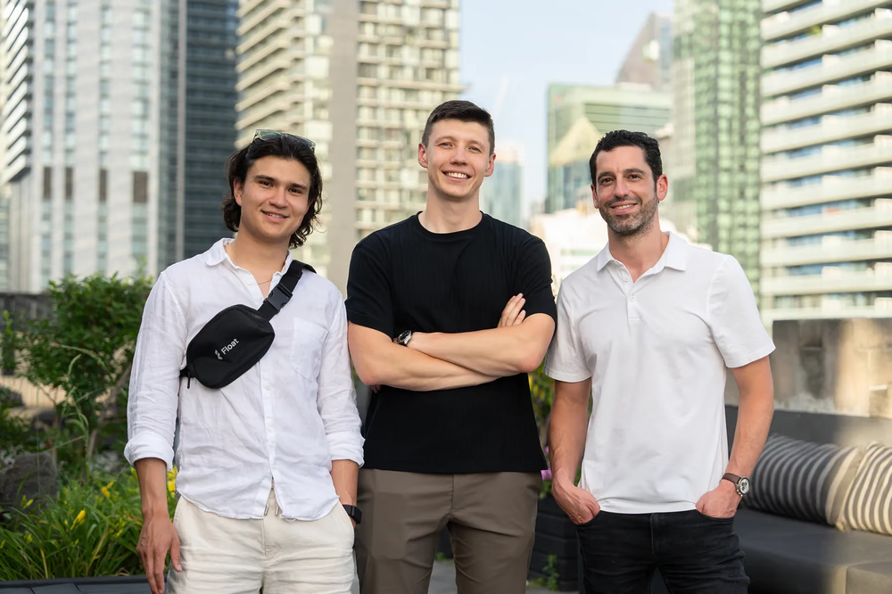 Three men (left man is wearing a white button-up shirt with a cross-bag, middle man is crossing his arms and wearing black shirt with grey pants, and the right man is wearing a white polo shirt) posing in front of skyscrapers