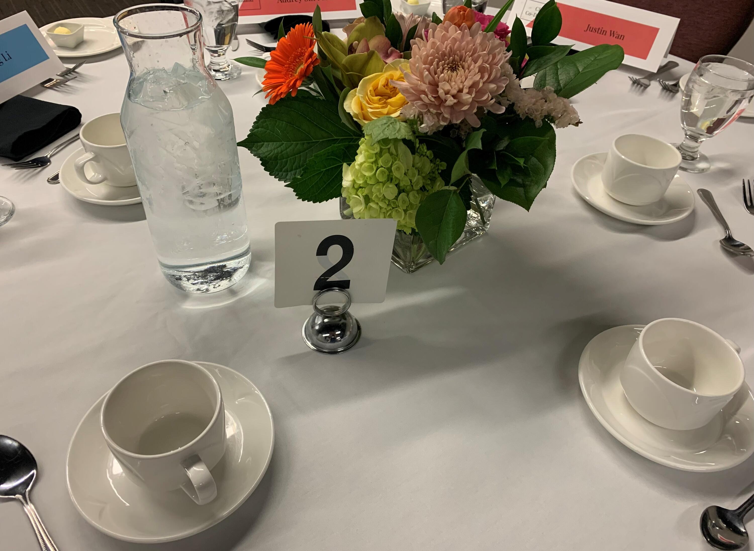 Table 2 at the CFM Graduation Dinner set with dishes, name cards, a pitcher of ice water, and a flower centrepiece