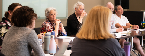 A woman listens intently to her workshop group as they discuss a scenario around a circle of tables. 