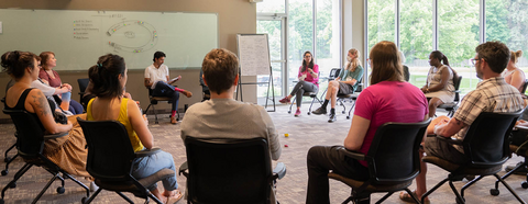 A group of workshop attendees sit in chairs in a circle to discuss scenarios and strategies. A large window brings light into the classroom.