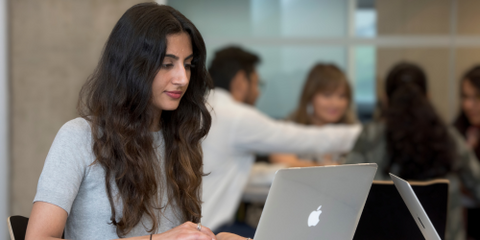 Person with long dark hair sitting at laptop computer. People in background.