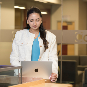 Person standing with laptop wearing white jacket and blue shirt.