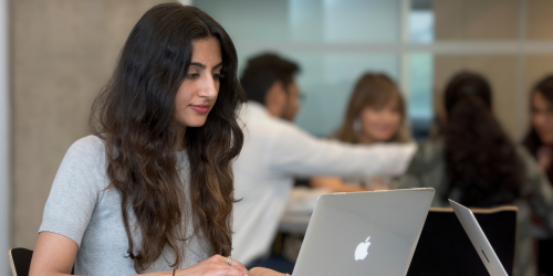 Person with long dark hair sitting at laptop computer. People in background.