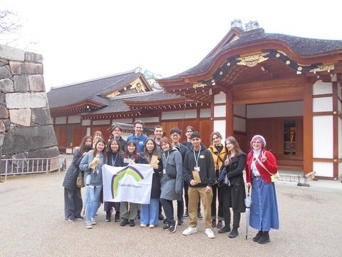 Students stand in front of a Japanese building holding a sign with the Kakehashi logo. 
