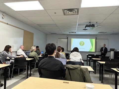 Brad Conrad stands at the front of the class next to a screen showing a powerpoint. Students are seated facing him at desks throughout the room. 