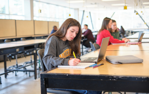 Student writing at a table with a notebook and a laptop