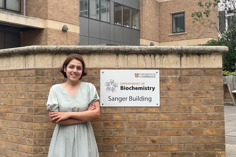 Atiyeh Ahmadi standing in front of a sign reading, "University of Cambridge: Department of Biochemistry - Sanger Building