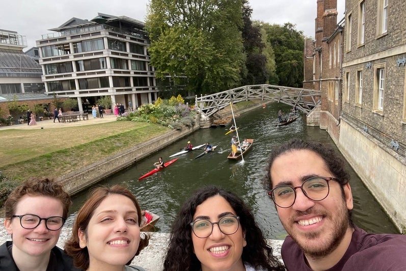 Four people take a selfie in front of a river where people are kayaking and canoeing.