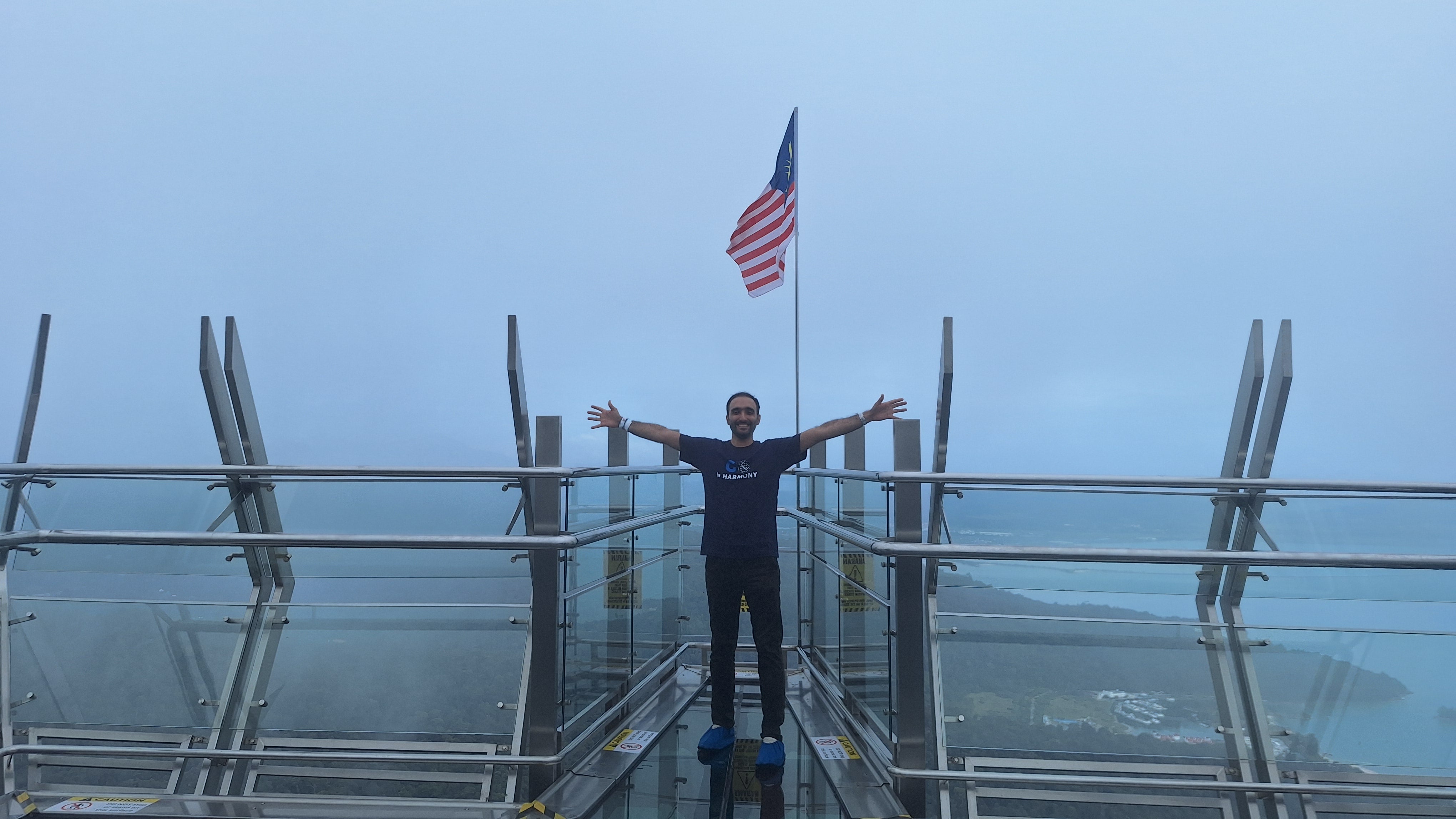 Mohammad Shojaati standing on a glass bridge with his arms out. There is a foggy landscape in the background.