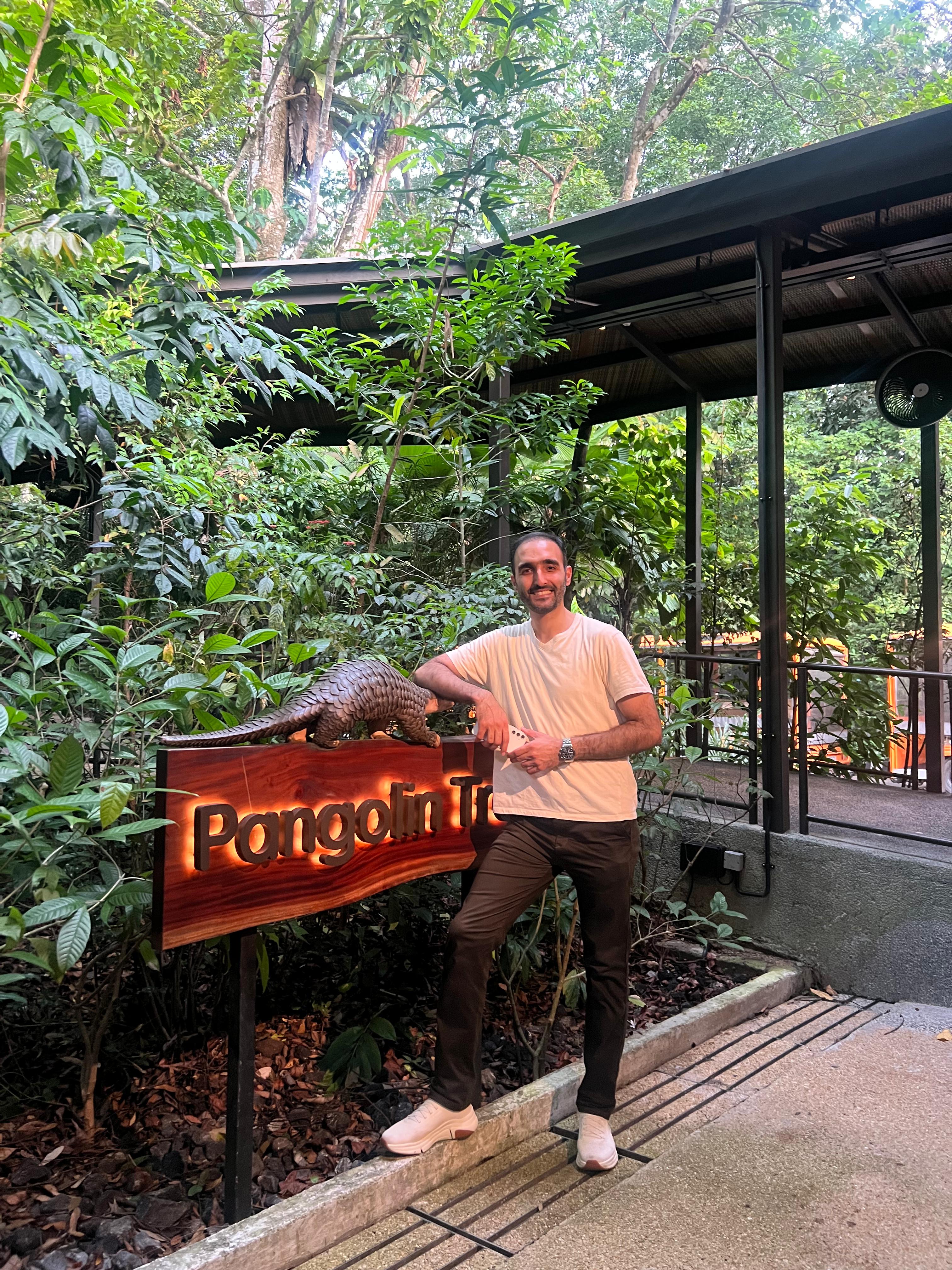 Mohammad Shoojati standing on a walkway in front of a jungle background. A sign he is leaning against reads, "Pangolin Trail"