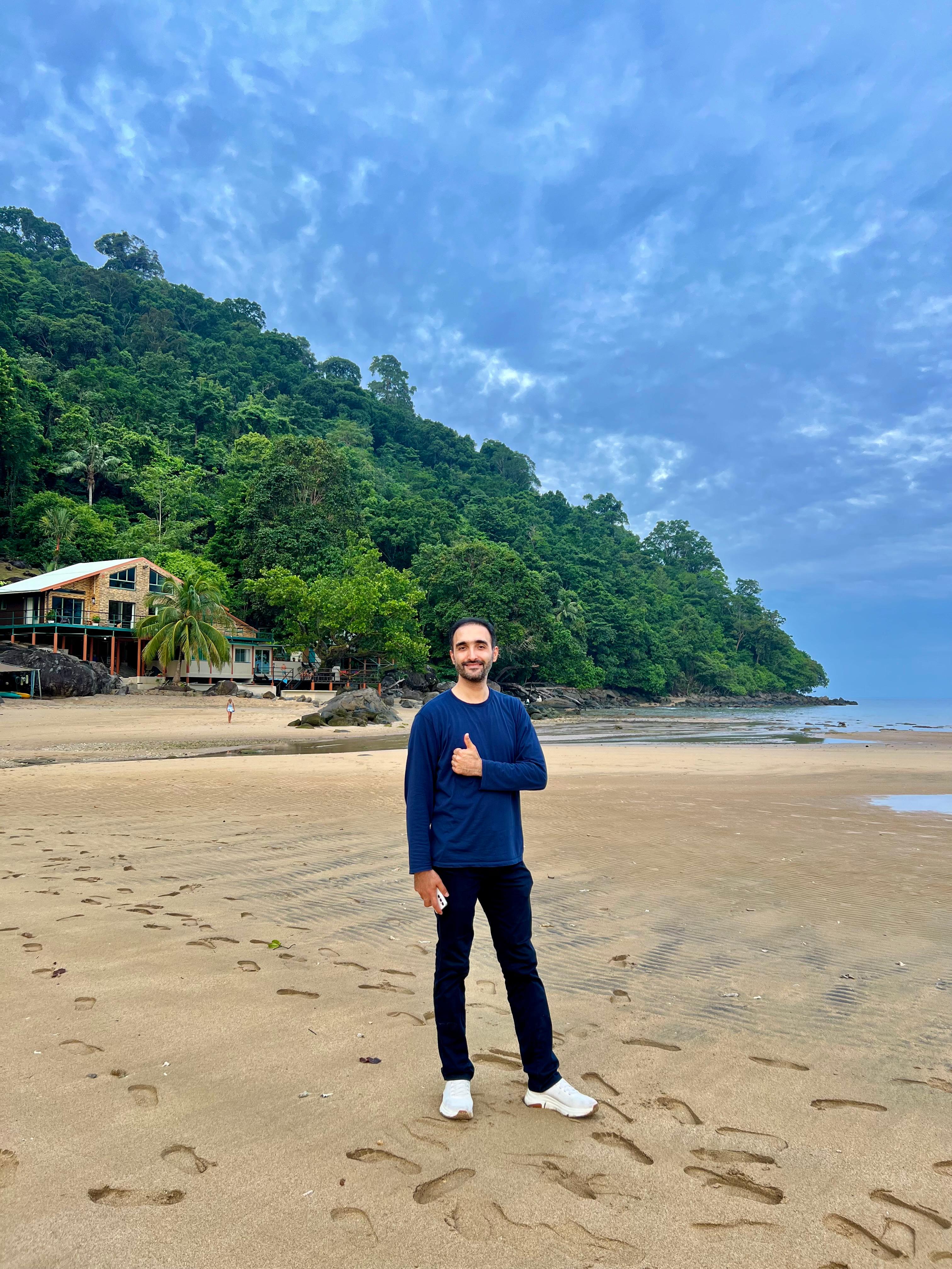 Mohammad Shojaati standing on a beach holding out a thumbs up. In the background there is a jungle, a body of water and a beach house.