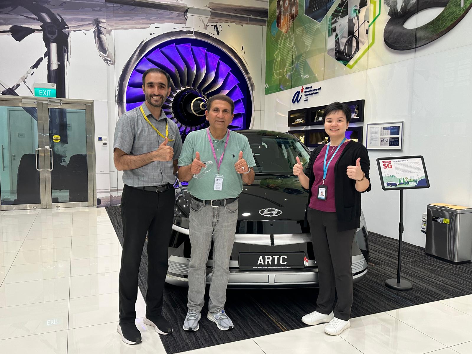 Three people standing in front of a display car holding out thumbs ups.