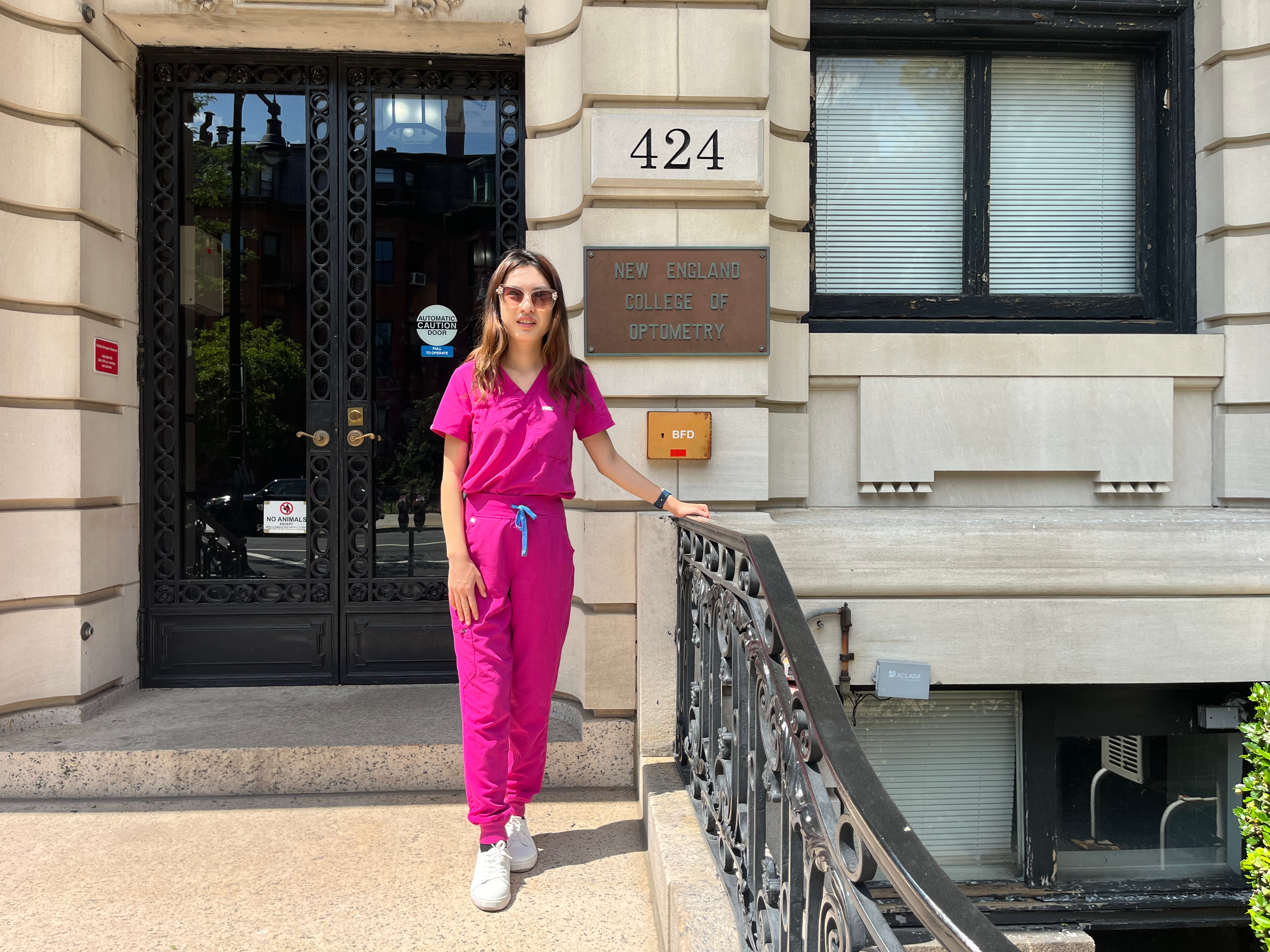 Sharon Qiu standing in front a building in pink scrubs. A plaque behind her reads: "New England College of Optometry"
