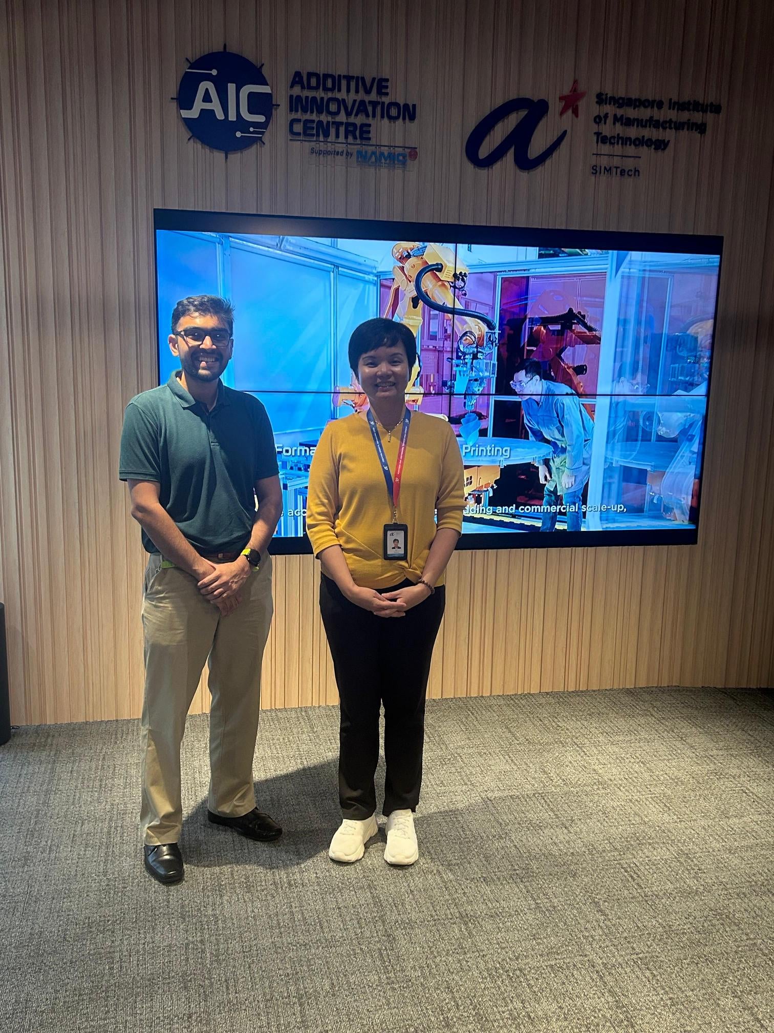 A man and a woman standing in front of a screen with conference lanyards around their necks.