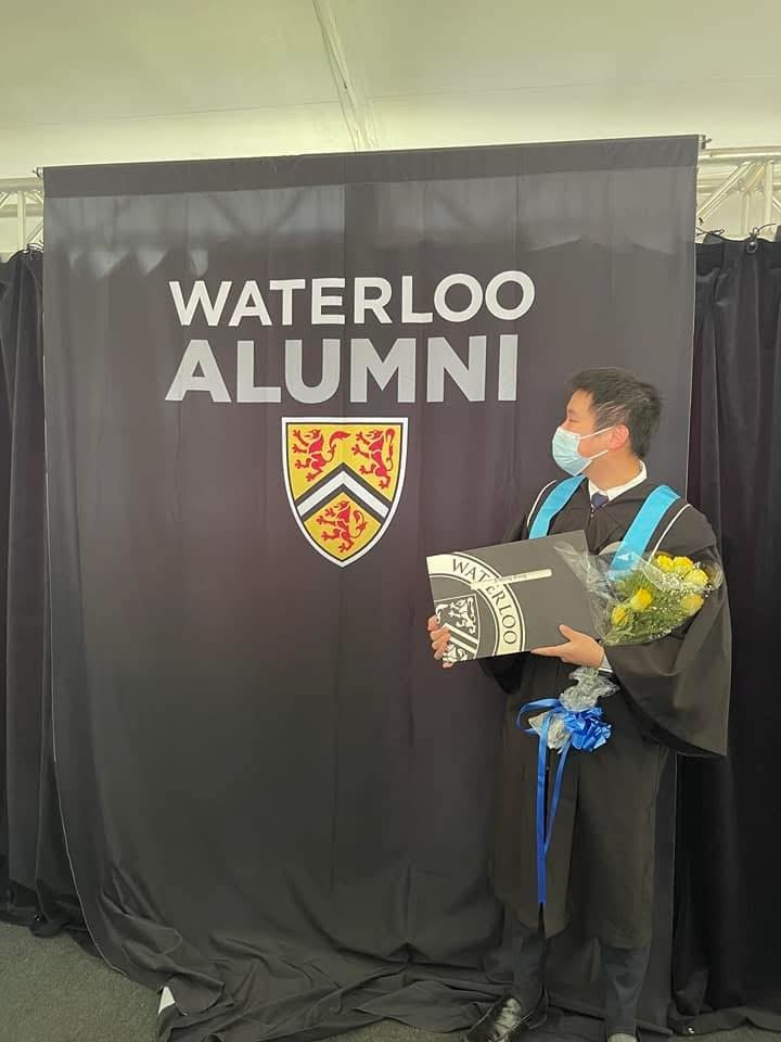 Graduate student Yueheng Song in graduate regalia standing in front of a photo backdrop that says "Waterloo Alumni"
