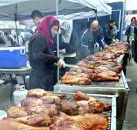 A volunteer cooks an impressive amount of turkey drumsticks on an open grill.