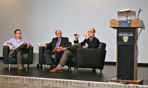 Three men sit in chairs on stage at a public lecture.