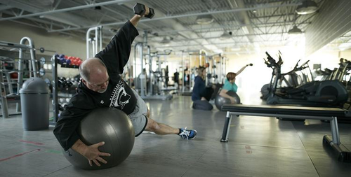 A man planks on an exercise ball while raising a dumbell with one arm.