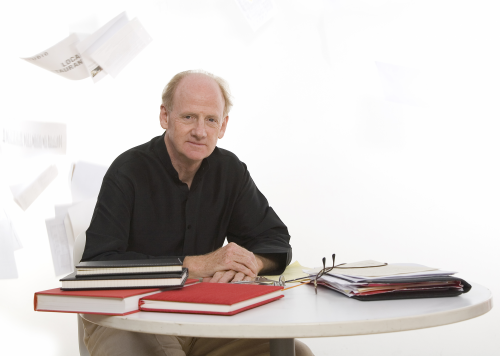 John Ralston Saul sits at a table with a sheaf of papers thrown in the air behind him.