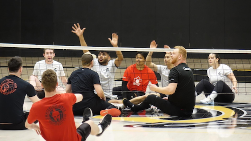 Athletes sitting on a floor with a volleyball net between them as part of a sitting volleyball game.