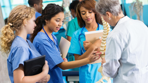 Students in medical scrubs look at a professor holding a model human spine.