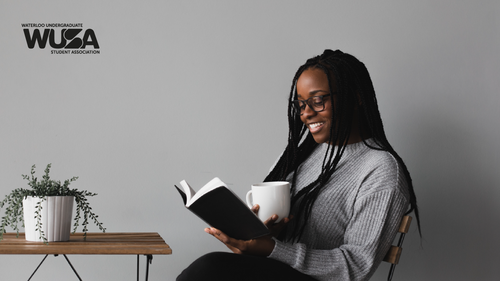 A woman with glasses sits with a mug at a table reading a book.