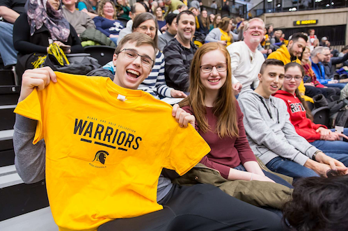 A smiling student holds up a Warriors t-shirt at a basketball game.