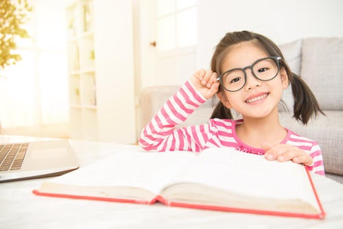 A child adjusts her glasses as she reads a book and smiles.