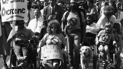 Disability activists, some walking with service animals, others in wheelchairs, march for accessibility with protest signs.
