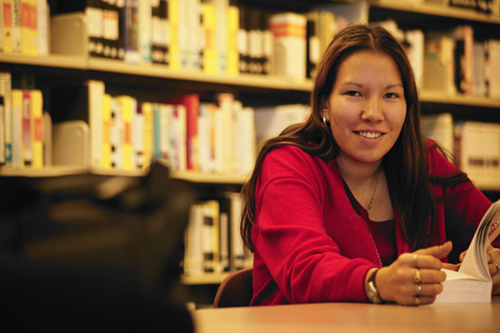An Indigenous woman sits at a table in a library with a book.