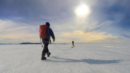 Two people in cold-weather gear walk across an ice field.