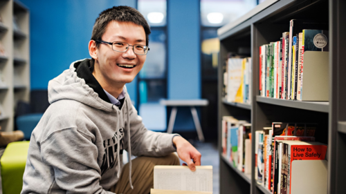 Co-op student Jun Chen Li smiles as he checks out books on a library shelf.