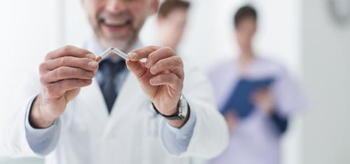 A man in a lab coat snaps a cigarette in two with his hands.