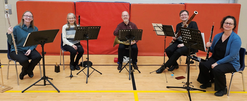 Five members of the wind ensemble sit with their instruments in a school gymnasium.