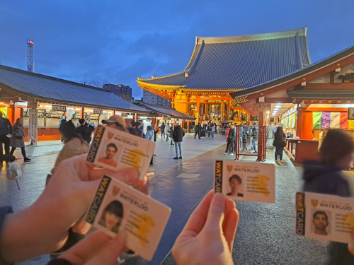 Students hold up their WatCards for a photo taken against the backdrop of traditional Japanese architecture.