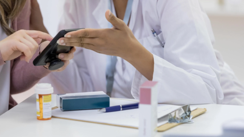 A pharmacist in a lab coat assists a patient look something up on their phone.