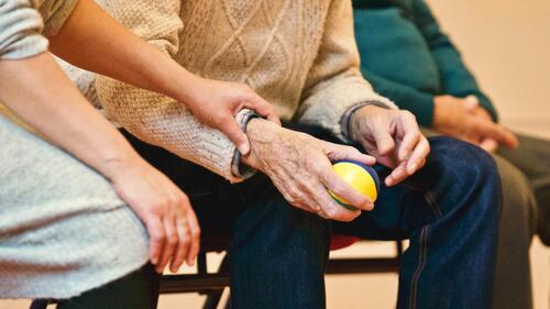 An elderly person holds a ball in their hands while a support worker gently holds the person's arm.