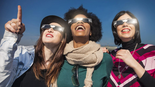 Three women wearing eclipse glasses look upward.
