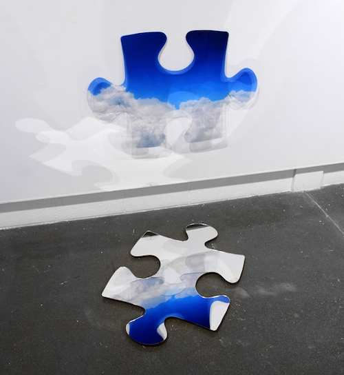 A large puzzle piece has fallen out of a wall onto the ground, revealing a blue sky and clouds beyond.