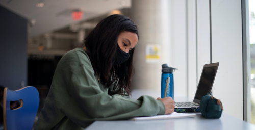 A young woman wearing a mask works at a laptop.