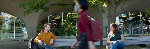 Students sit physically distant on a bench while a masked person walks nearby.