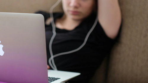 A young woman wearing headphones stares at her laptop screen.