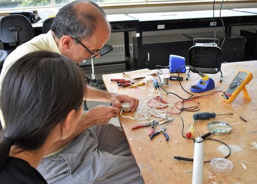 A man works on repairing an electrical device, with a soldering iron nearby, while a person watches.