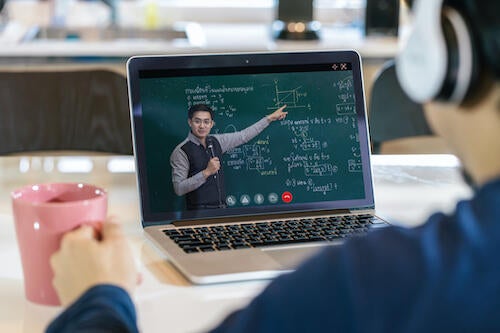 A student watches a teacher conduct a class on a laptop screen.