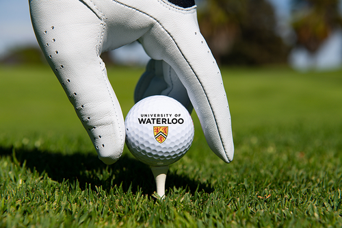 A hand sheathed in a golfer's glove places a UW-branded golf ball on a tee.