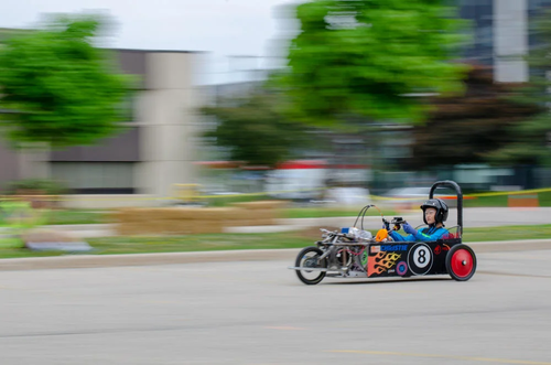 An action shot of a student driving an electric go-kart-style racecar near University buildings.