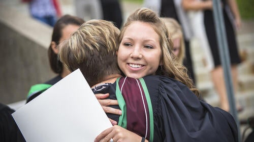 Two graduates in convocation robes hug.
