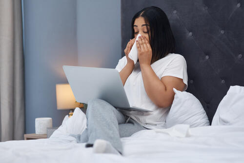 A woman blows her nose with a kleenex while sitting in bed.